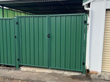 Green corrugated double gates hang beside a white garage wall opening onto asphalt with a metal carport roof behind.