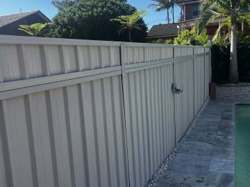 Off-white corrugated panels and a latched gate line a pebble strip next to stone pool pavers and palm trees.