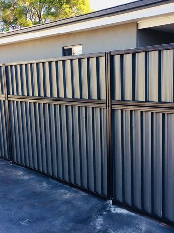 Monument panels with a shorter upper infill run along a dark concrete driveway beside a grey house.