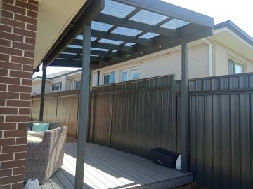 The scene shows a mid grey corrugated steel fence where an upper infill raises the height, concrete paving runs alongside.