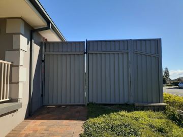 Dark grey pedestrian gate and tapered sheet panels sit on brick paving beside a modern grey and white house.