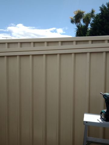Matching cream panels carry an upper corrugated infill with a ladder and drill resting against the fence under palm trees.