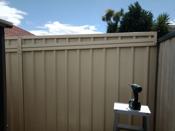 Matching cream panels carry an upper corrugated infill with a ladder and drill resting against the fence under palm trees.