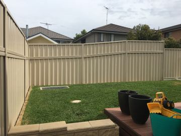 The scene shows a mid grey corrugated steel fence where an upper infill raises the height, sleepers form the base.