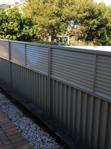 The scene shows a mid grey corrugated steel fence where an upper infill raises the height, residential buildings appear beyond the line.