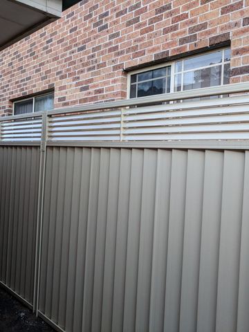The scene shows a mid grey corrugated steel fence where concrete paving runs alongside.