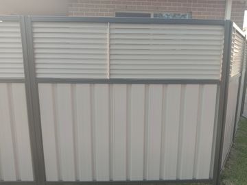 The scene shows a light grey corrugated steel fence where concrete paving runs alongside.