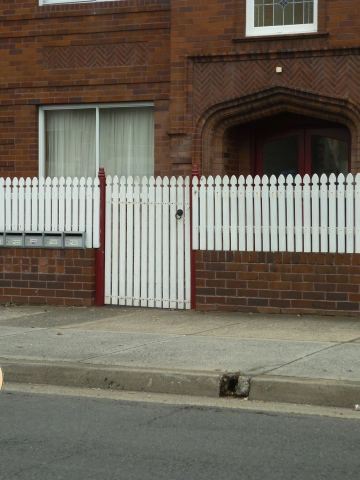 The scene shows a mid grey corrugated steel fence where concrete paving runs alongside.