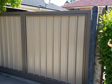 The scene shows a light grey corrugated steel fence where concrete paving runs alongside.