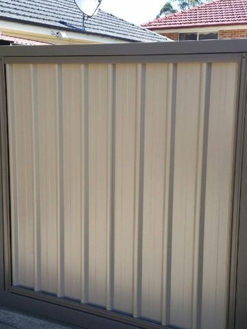 The scene shows a light grey corrugated steel fence where concrete paving runs alongside.