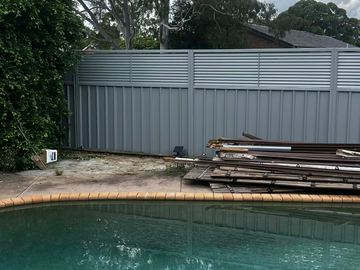 Grey panels with louvre tops border pool coping beside a tall hedge and stacked old timber fence materials.