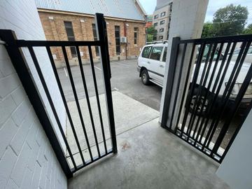 Black flat-top picket gate and panel open from a white brick porch toward an aggregate path and parked cars.