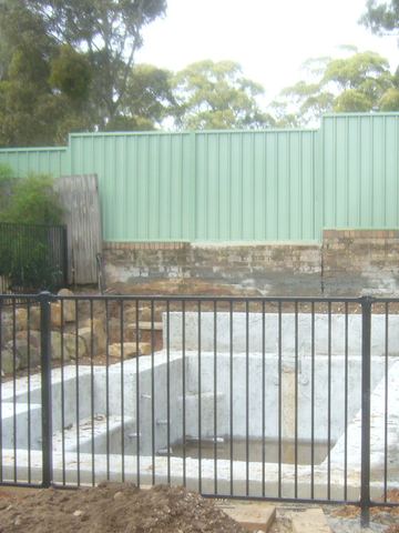 Black tubular panels ring a concrete pool shell amid green stepped Colorbond, cream-red side fencing and rockery.