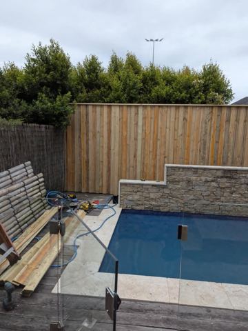 Vertical timber slats on stone tiers stand beside frameless glass pool fencing, tools and brushwood screening.