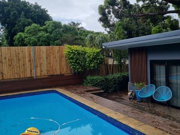 Natural pine palings on a dark sleeper base border a blue pool with timber deck and grey pool studio behind.