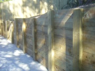 Stacked pine sleepers with front posts retain soil beside a light concrete path with scattered leaves.
