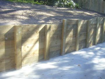 Horizontal sleepers anchored by posts wrap a corner where concrete paving meets a bare soil embankment and timber paling beyond.
