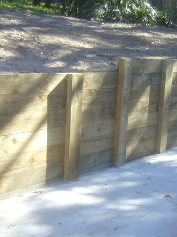 Horizontal sleepers anchored by posts wrap a corner where concrete paving meets a bare soil embankment and timber paling beyond.