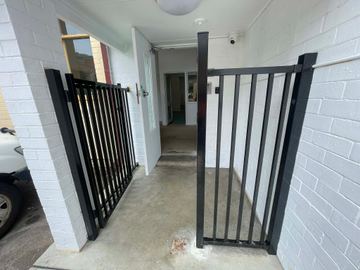 Black picket double gates stand open in a white brick entry with keypad and camera above a concrete floor.