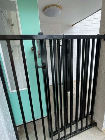 Black picket gate closes a white-brick alcove in front of a mint-green glazed door under a ceiling light.