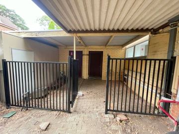 Black picket fence and gate span a brick-paved passage beneath a corrugated awning beside a cream brick building.